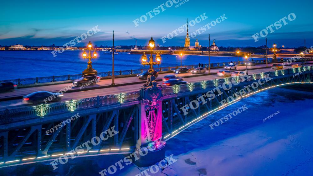 На фотографии изображён Троицкий мост, один из красивейших мостов Санкт-Петербурга, в вечерней подсветке. Заснеженная Нева и огни мостовых фонарей создают атмосферу волшебства зимней ночи. Вдали можно увидеть Петропавловскую крепость, символ исторического Петербурга, освещённую и выделяющуюся на фоне глубокого синего неба. Этот вид подчёркивает гармонию архитектуры и городской ночной жизни в холодное время года.